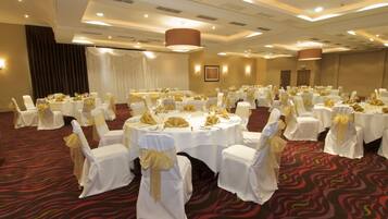 Un salón de banquetes con mesas redondas, sillas blancas con cintas doradas y una alfombra roja con estampado.