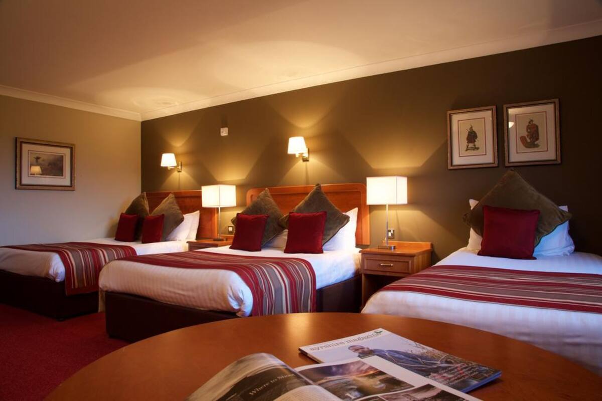 Quarto de hotel com três camas, cada uma com cabeceira, luminárias de cabeceira e almofadas. Há uma mesa redonda com uma revista no centro do quarto.