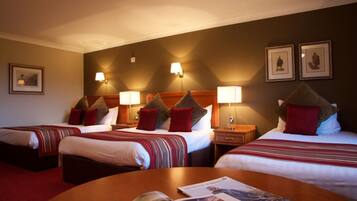 Habitación de hotel con tres camas, cada una con cabecero, lámparas de noche y almohadas. Hay una mesa redonda con una revista en el centro de la habitación.