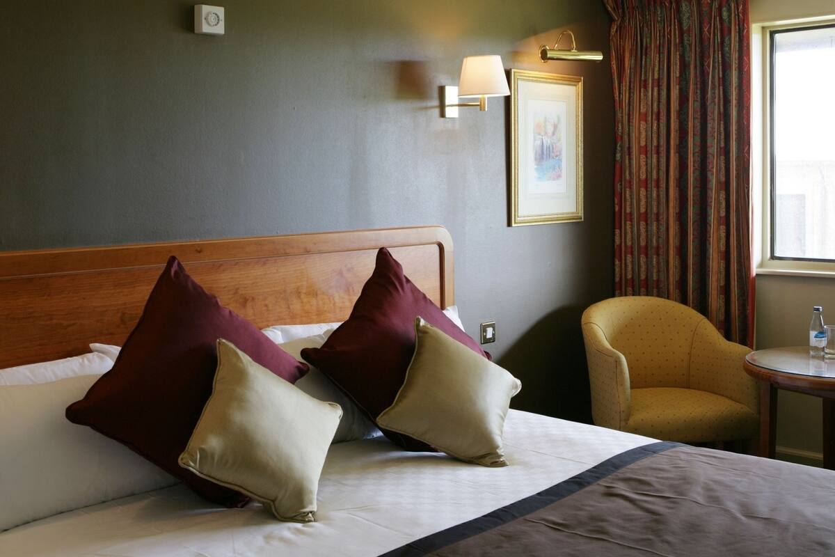 Quarto de hotel com uma cama, duas almofadas, uma cadeira, uma mesa, um abajur, um quadro e uma janela com cortinas.
