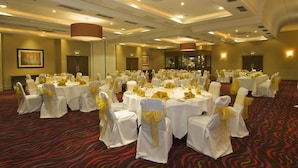 Un salón de banquetes con mesas redondas cubiertas con manteles blancos y sillas con cintas amarillas, dispuesto en una sala amplia con alfombra estampada e iluminación empotrada.