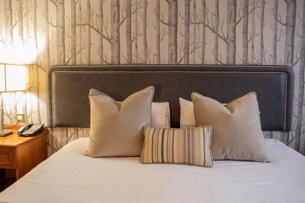 Uma cama bem arrumada com uma cabeceira escura, duas almofadas grandes e uma almofada decorativa estampada. É visível uma mesa de cabeceira com um abajur e um telefone.