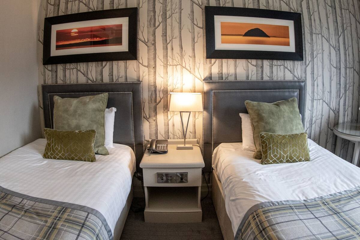 Quarto de hotel com duas camas, uma mesa de cabeceira com abajur e duas fotografias emolduradas na parede.