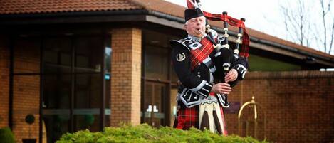 A person in traditional Scottish attire playing bagpipes in front of a building with a brick facade and large windows.