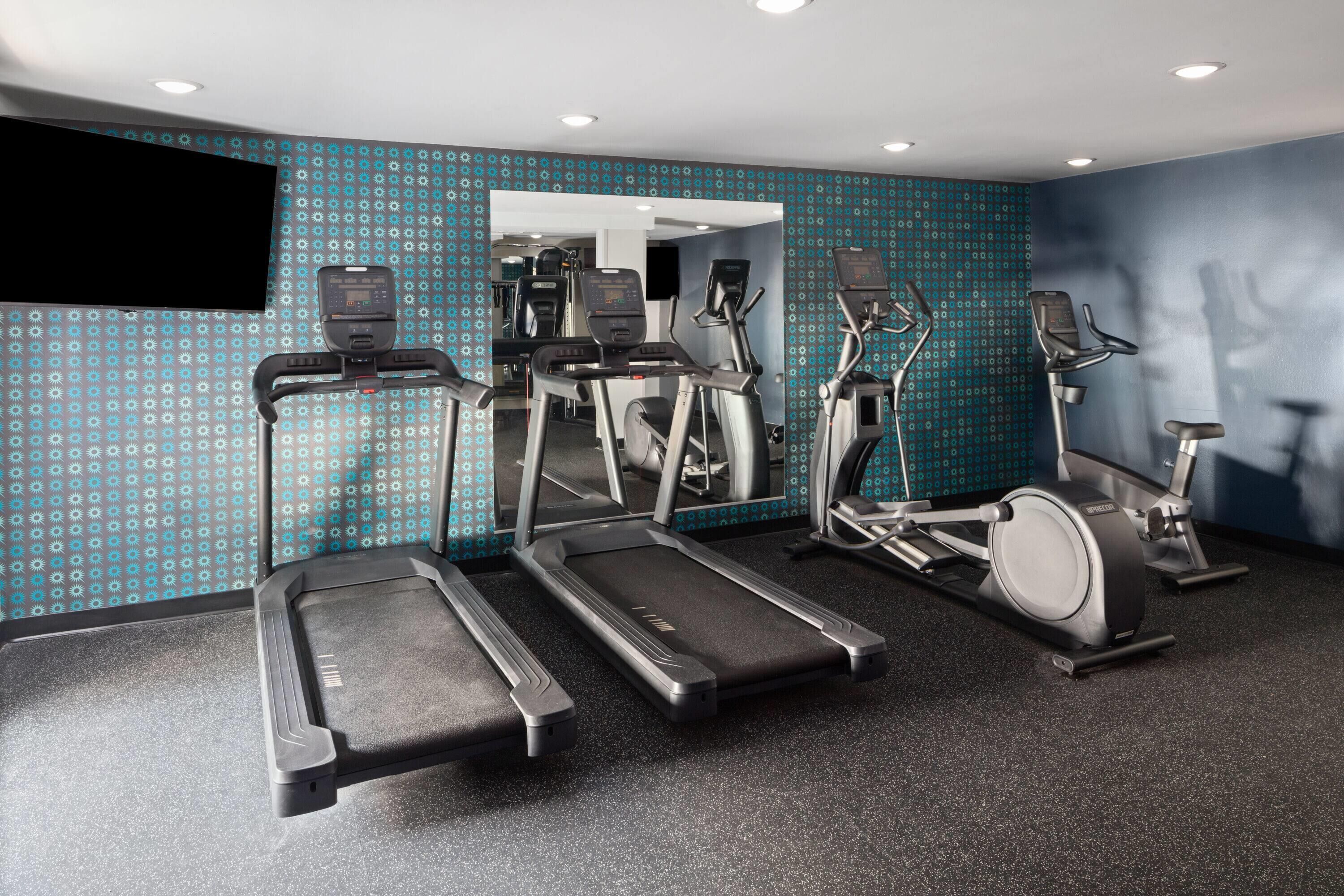 A gym with treadmills, elliptical machines, and a TV screen on the wall.