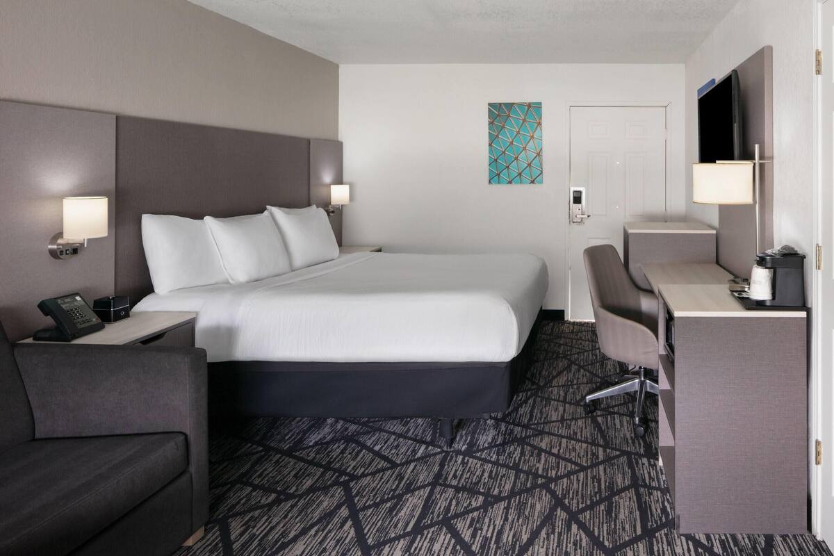 A hotel room with a bed, a desk, a chair, a phone, and a TV.