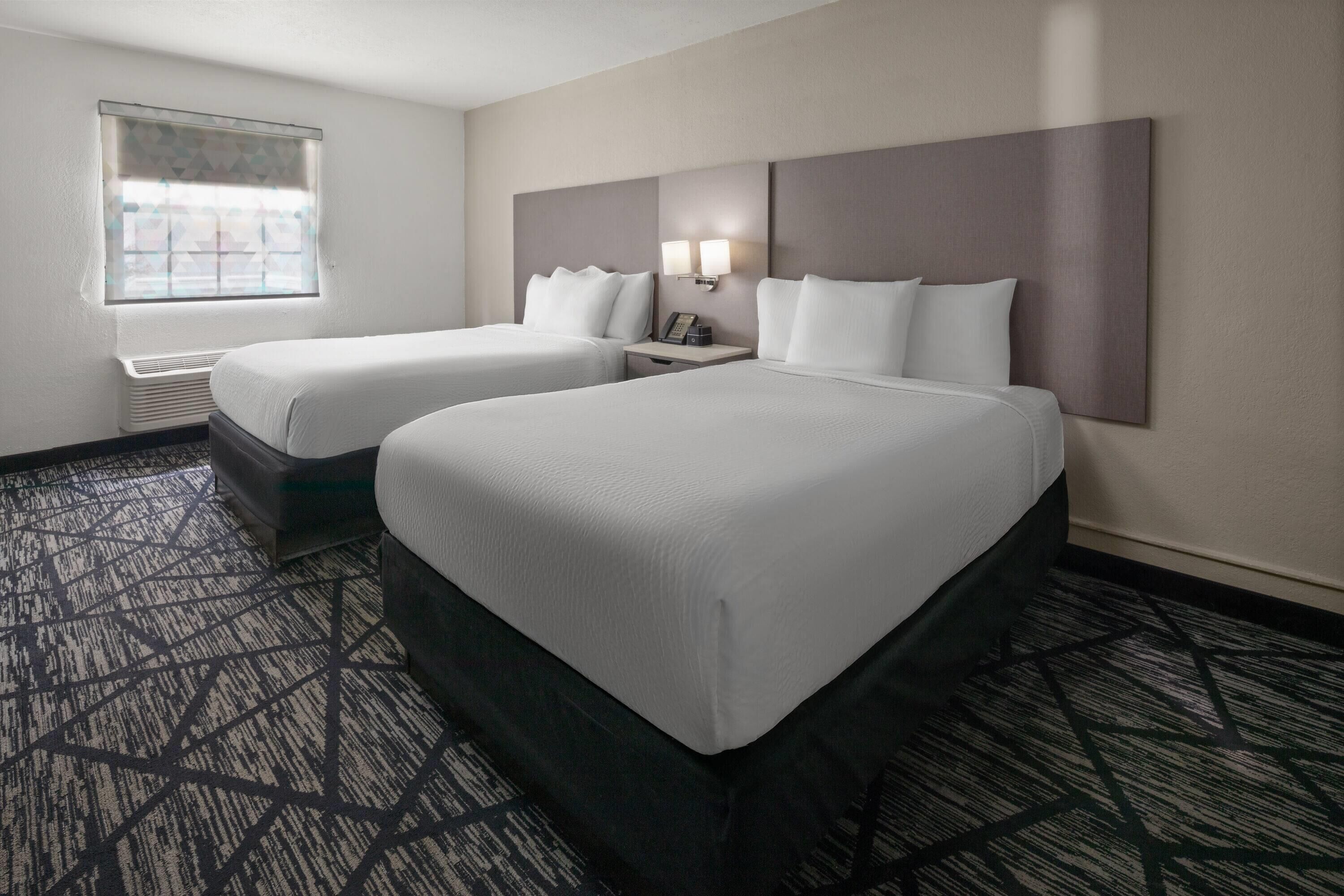 A hotel room with two beds, a window with blinds, a wall-mounted light, and a patterned carpet.