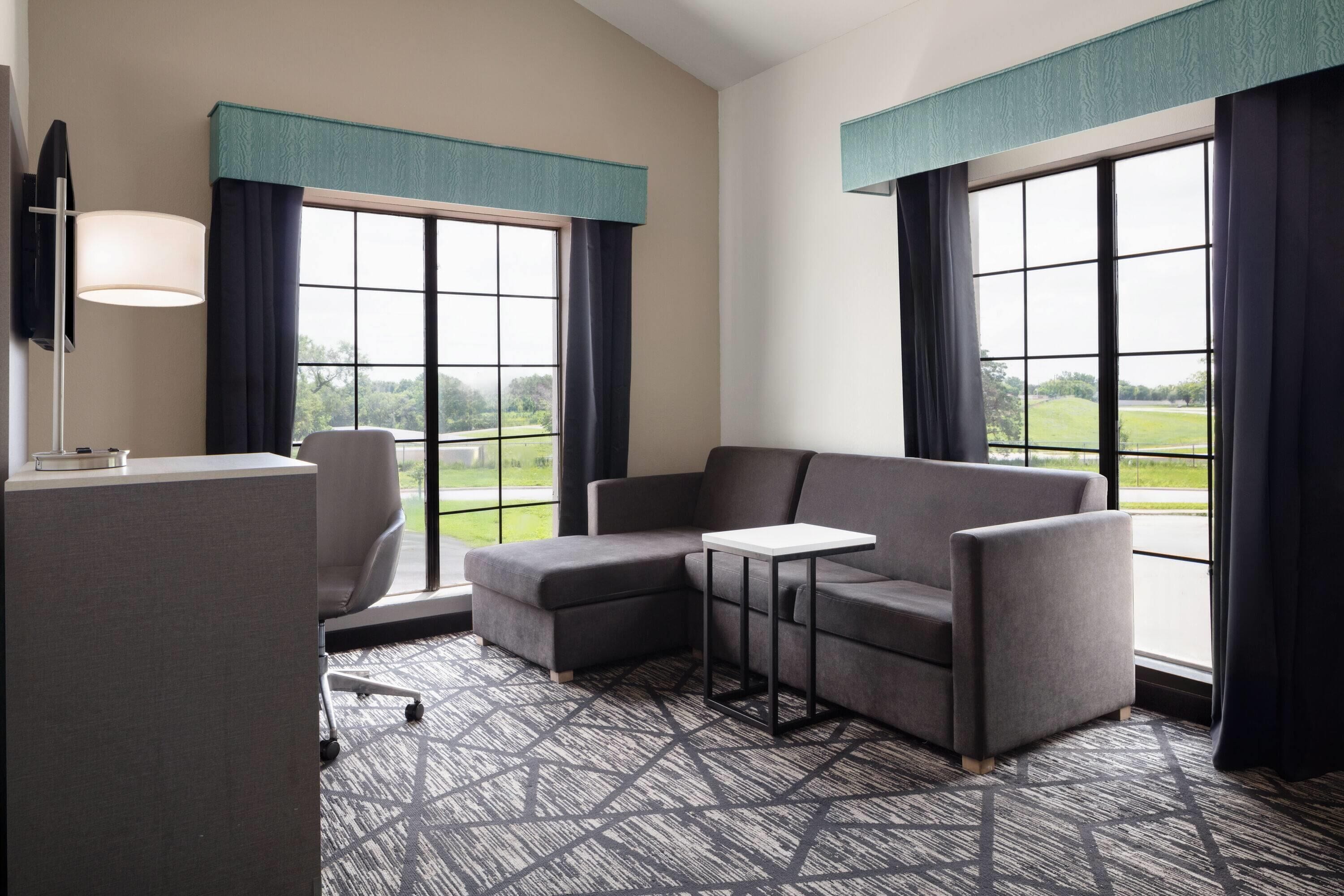 A hotel room with a sectional sofa, a small table, a desk, and a TV.