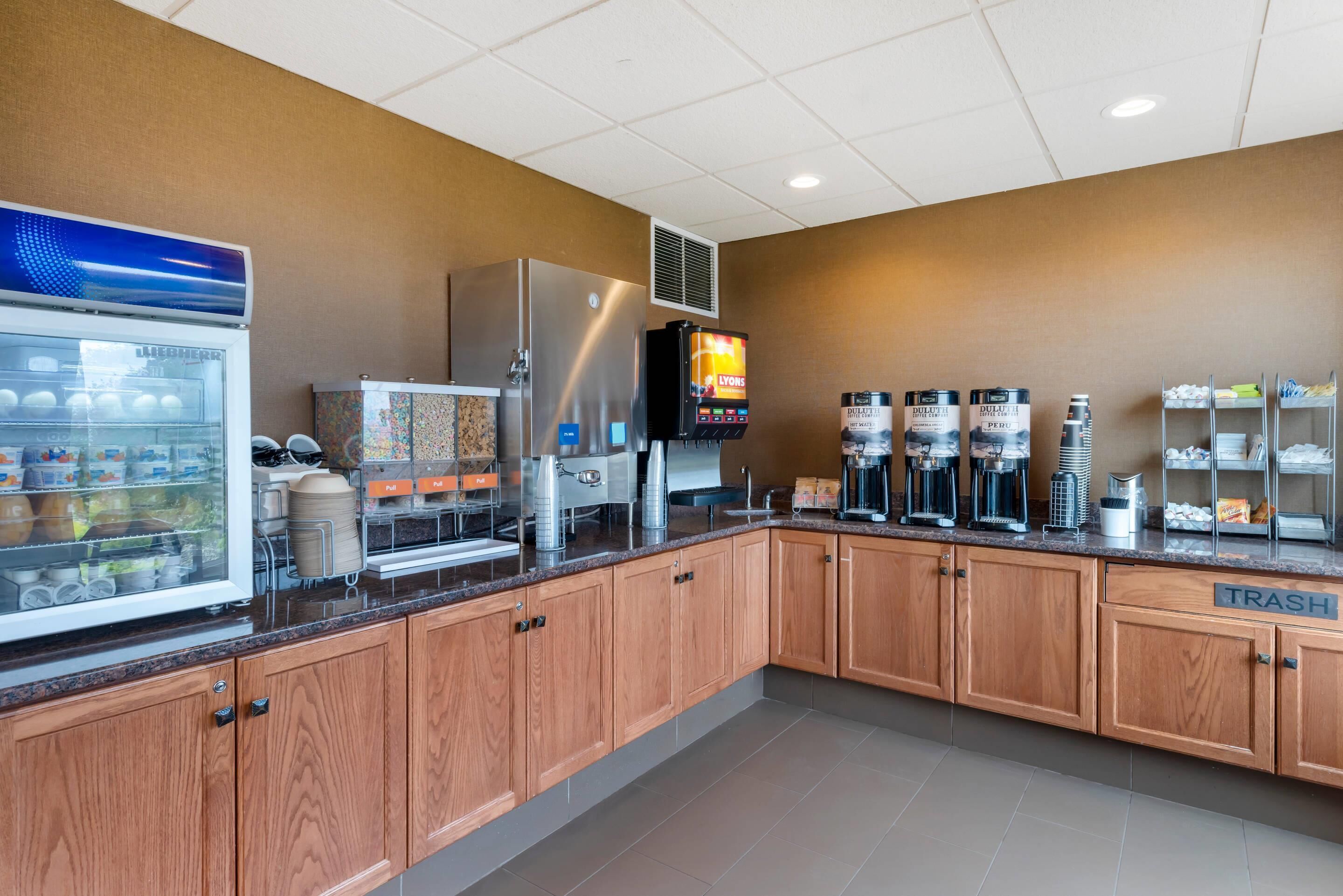 A self-service breakfast area with a refrigerated display, coffee machines, and a trash bin.