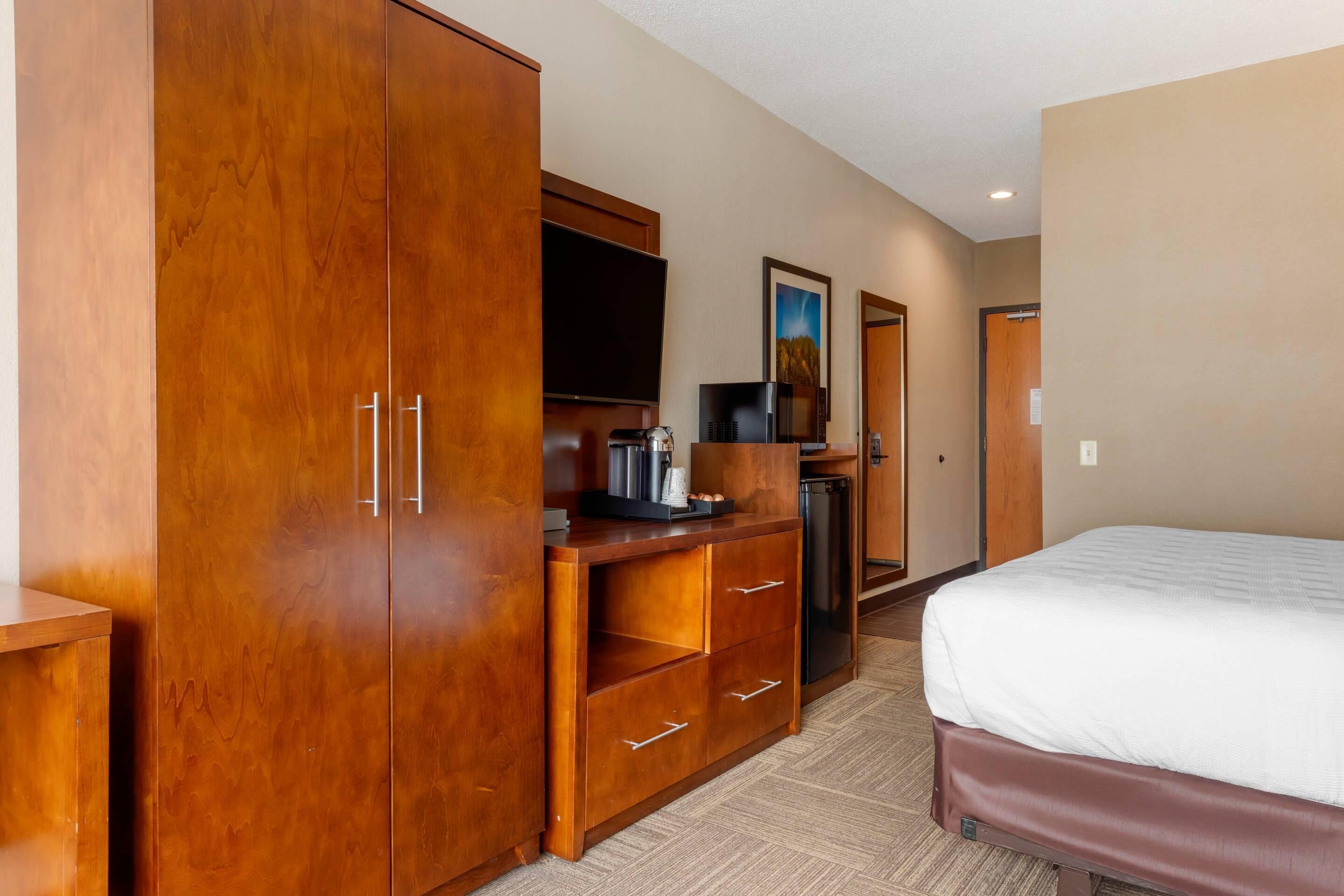 A hotel room with a large wooden wardrobe, a flat-screen TV, a microwave, a coffee maker, and a bed with a white mattress.