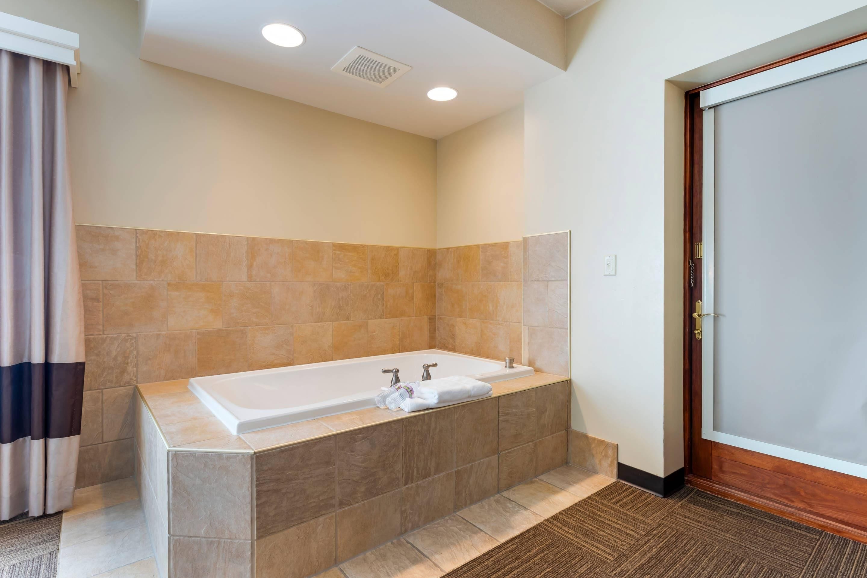 A modern bathroom with a large bathtub, beige tiles, and a wooden door.