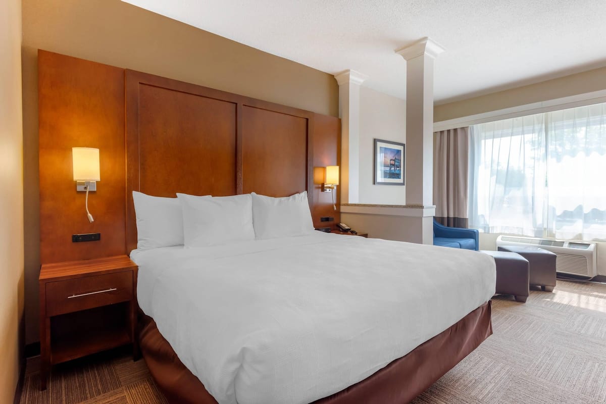 A hotel room with a large bed, wooden headboard, bedside lamps, a nightstand, and a view of the city through large windows.