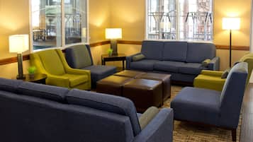 A hotel lobby with a large grey sofa, a yellow armchair, and a brown ottoman. There are two floor lamps and a small table with a plant.
