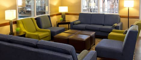 A hotel lobby with a large grey sofa, a yellow armchair, and a brown ottoman. There are two floor lamps and a small table with a plant.