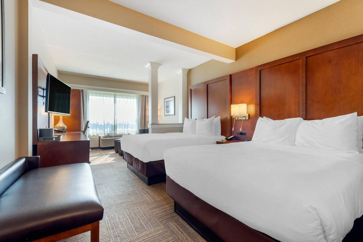 A hotel room with two beds, a desk, a TV, and a view of the city.