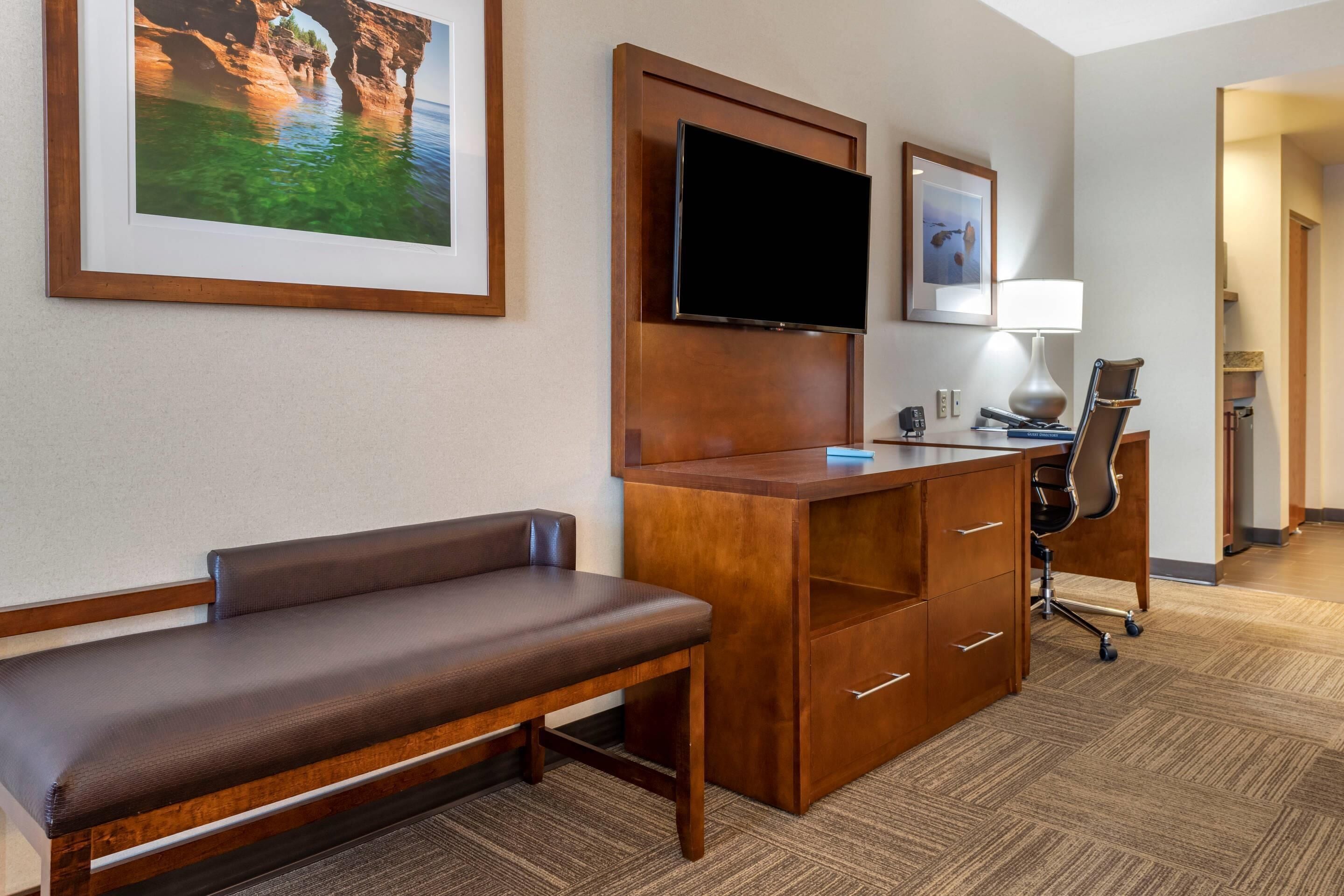 Hotelkamer met een houten bureau, een flatscreen televisie, een bankje, een bureaulamp en een lijst met een kustlandschap.
