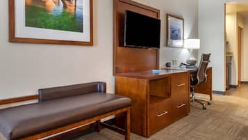 Hotelkamer met een houten bureau, een flatscreen televisie, een bankje, een bureaulamp en een lijst met een kustlandschap.
