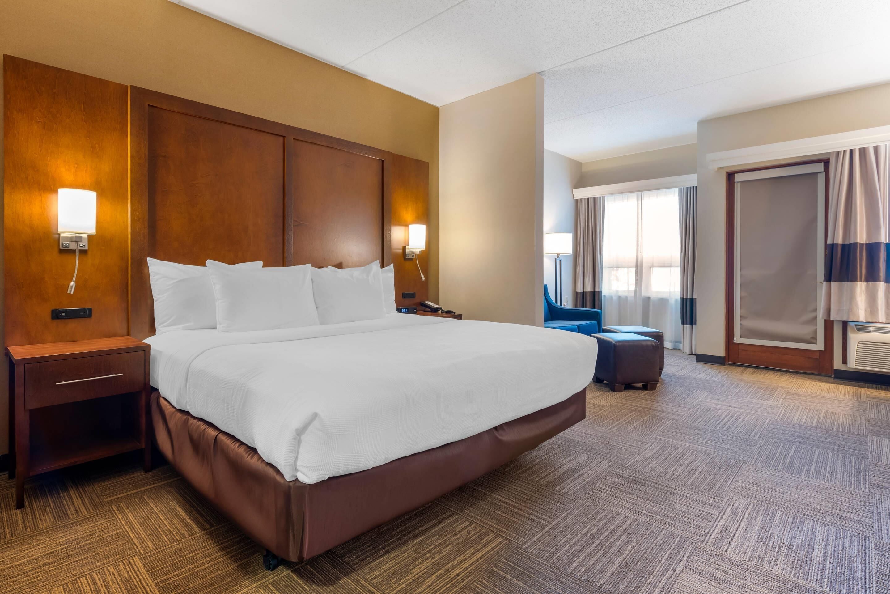 A hotel room with a large bed, wooden headboard, two bedside tables, a nightstand, a small seating area, and a large window with curtains.