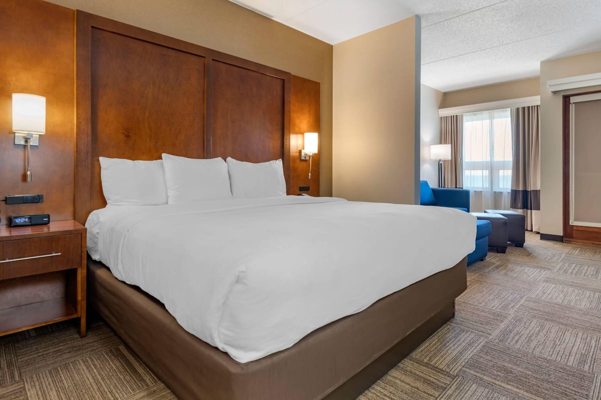 A hotel room with a large bed, wooden headboard, bedside lamps, a nightstand, and a blue armchair.