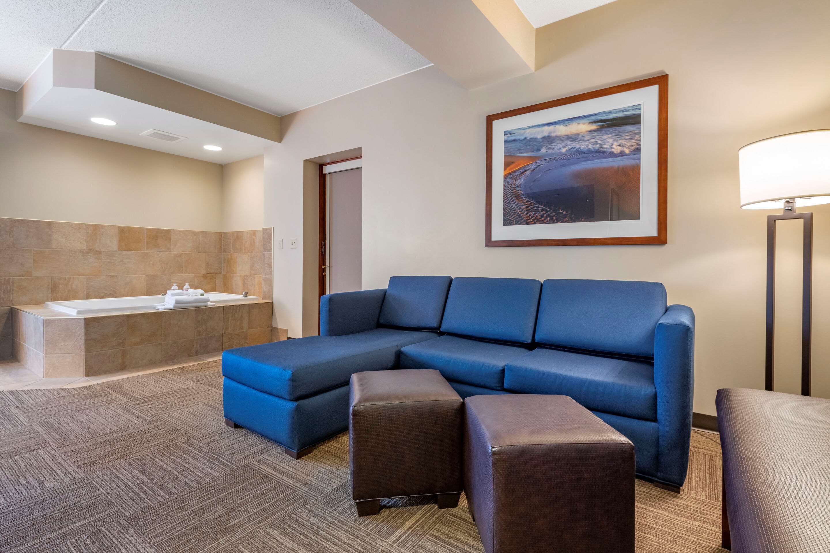 A modern hotel room with a large sectional sofa, a small ottoman, a bathtub, and a framed picture on the wall.