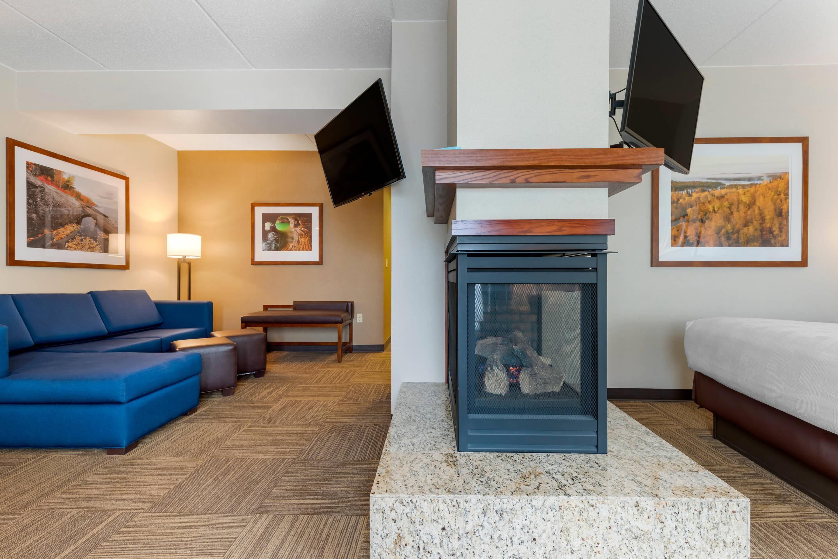 Quarto de hotel com sofá modular azul, uma mesinha, um banco, lareira e duas televisões na parede.