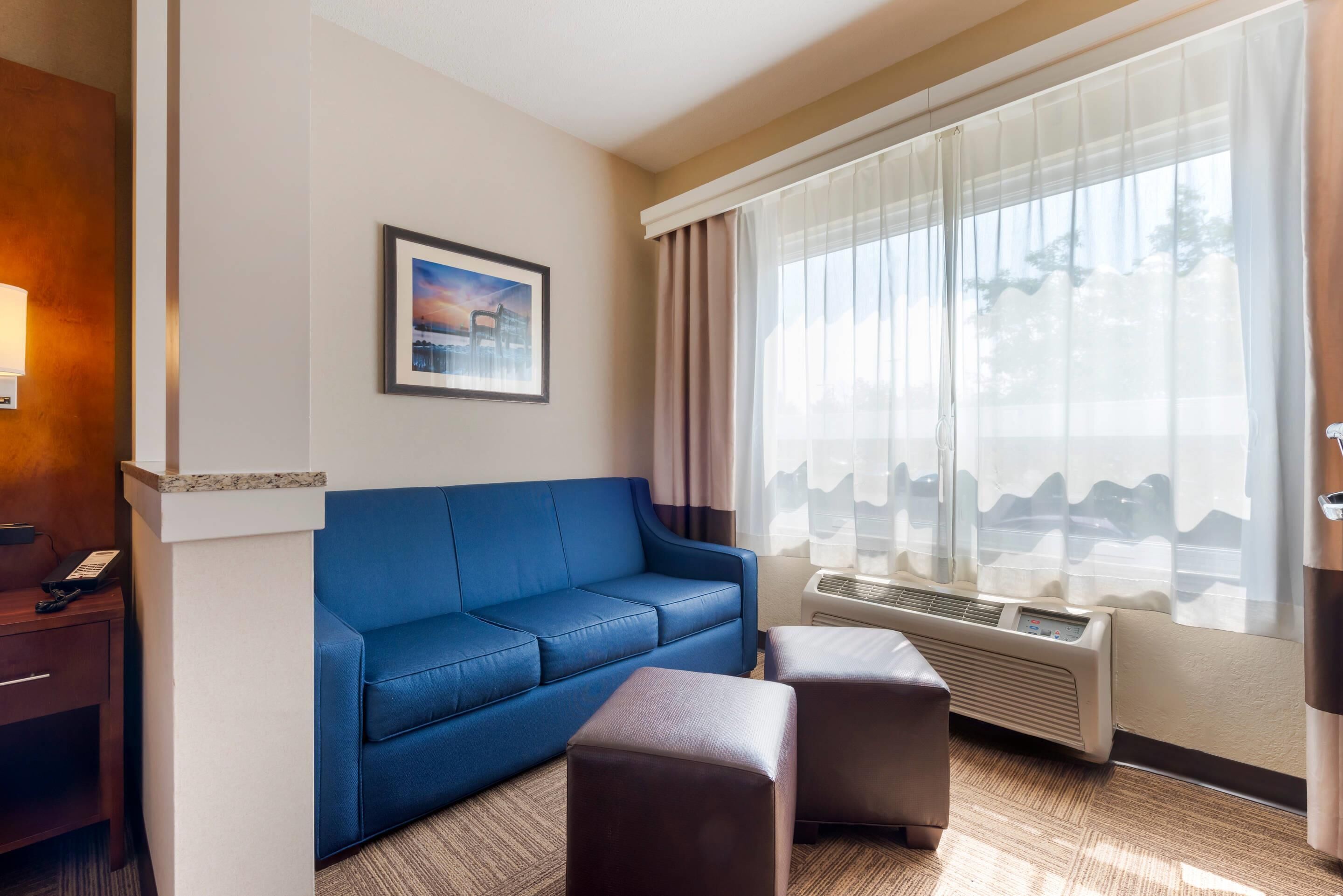 Habitación de hotel con sofá azul, un pequeño reposapiés, un cuadro enmarcado en la pared y un ventanal con cortinas.