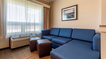 Une chambre d’hôtel avec un canapé d’angle bleu, un appareil de climatisation, une fenêtre avec des rideaux légers et un tableau encadré accroché au mur.