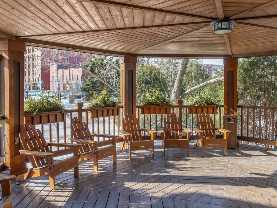 A wooden deck with Adirondack chairs, a wooden railing, and a view of buildings and trees.