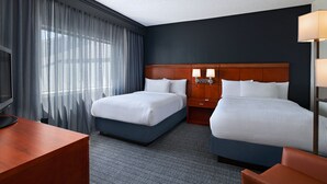 A hotel room with two beds, a desk, a TV, and a large window with curtains.