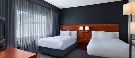 A hotel room with two beds, a desk, a TV, and a large window with curtains.