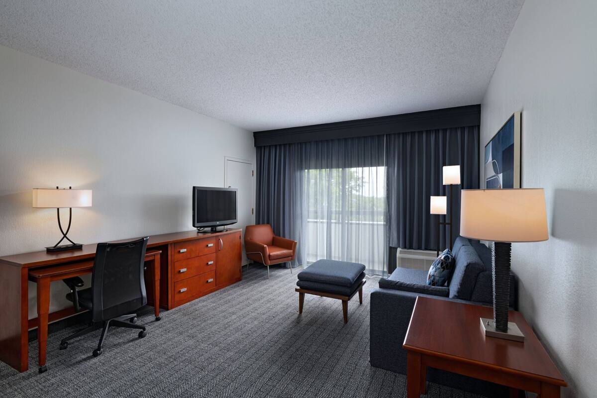 Habitación de hotel con escritorio de madera, televisor, sillón de cuero, sofá y lámpara.