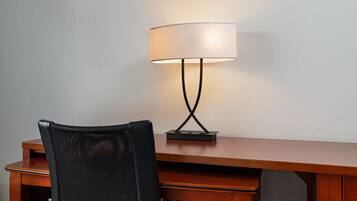 A wooden desk with a black mesh chair and a table lamp with a white shade.