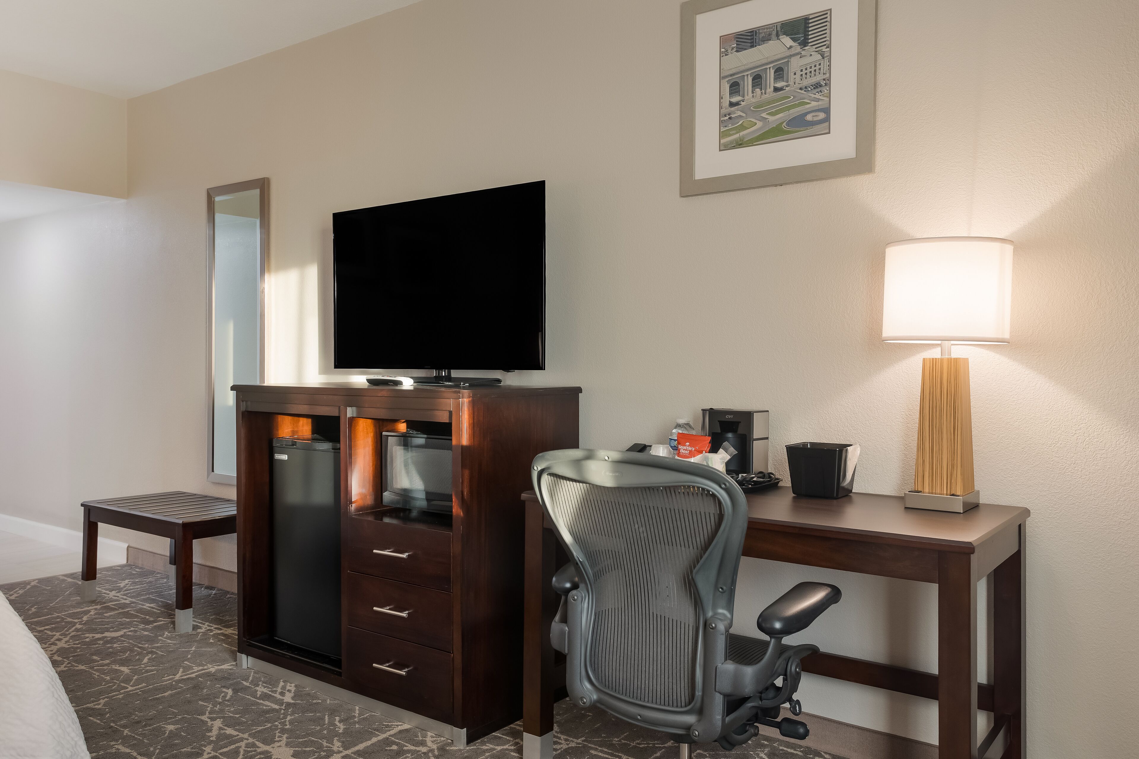 Habitación de hotel con cama, escritorio y silla, televisor, cafetera, lámpara y un cuadro en la pared.
