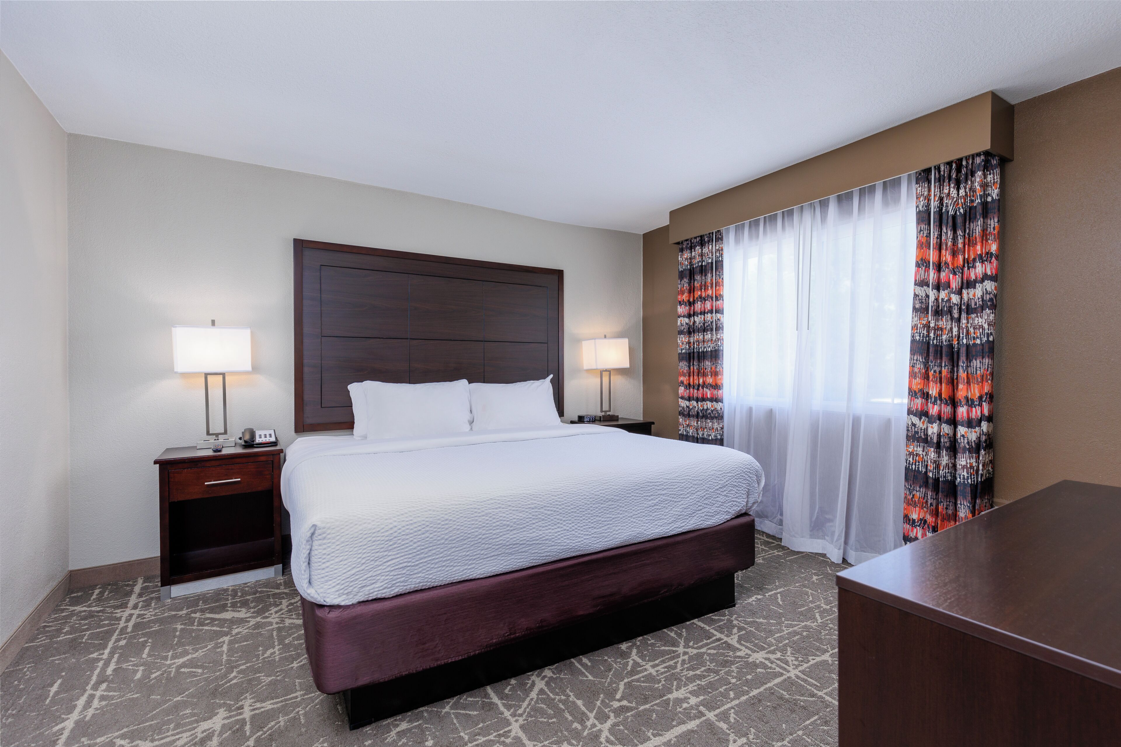 A hotel room with a large bed, two bedside tables with lamps, a wooden headboard, and a patterned curtain.