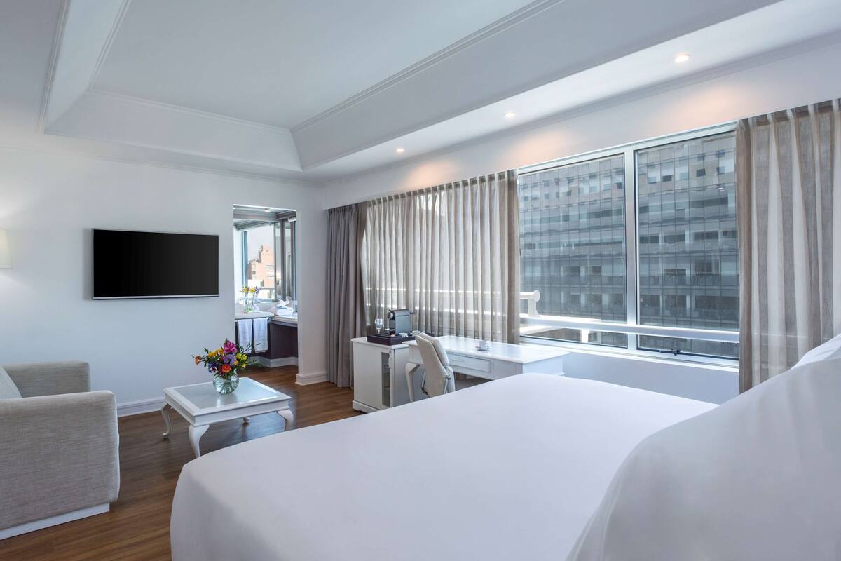 A modern hotel room with a large bed, a desk with a computer, a chair, a TV mounted on the wall, and a view of the city through large windows.