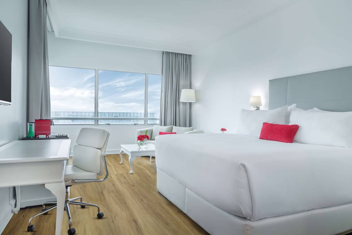 A hotel room with a large bed, a desk, a chair, a small table with a vase of flowers, and a view of the sky through large windows.