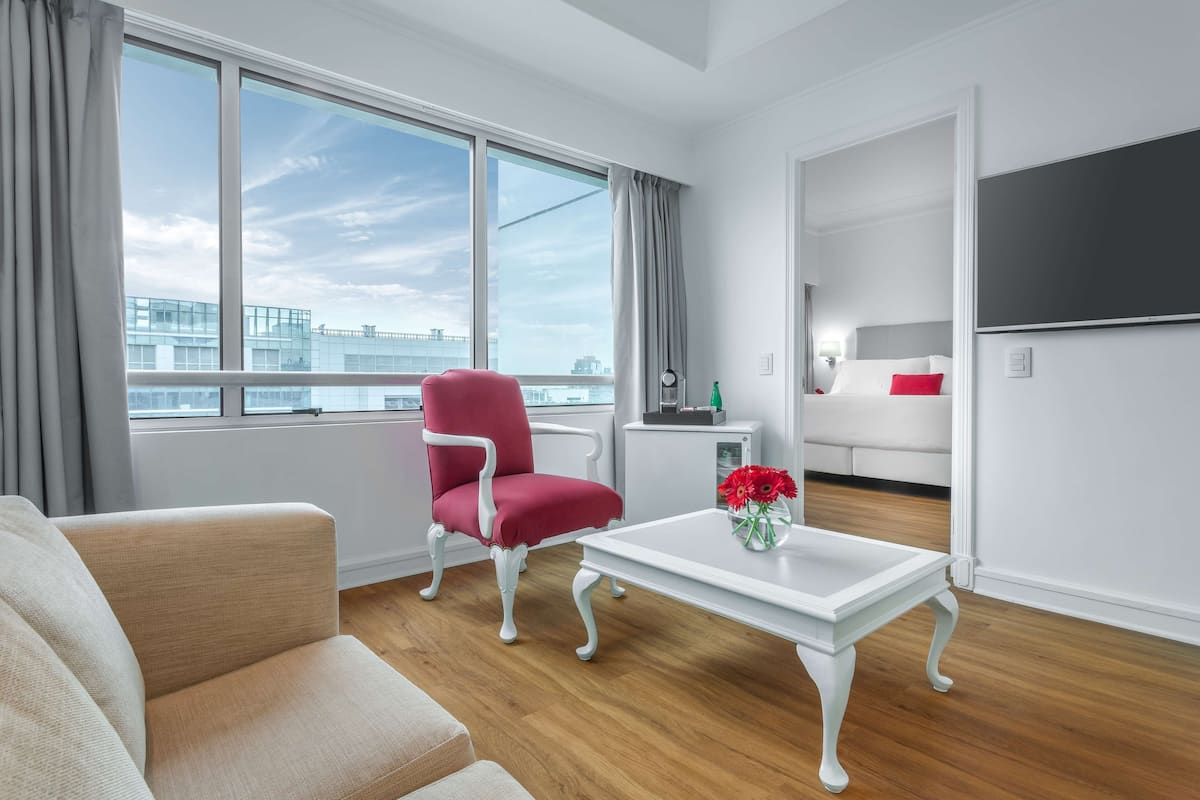 A modern hotel room with a large window, a red armchair, a white sofa, a small white table with a vase of flowers, and a bed with white bedding and a red pillow.