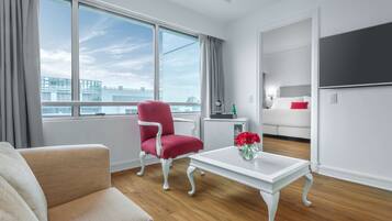A modern hotel room with a large window, a red armchair, a white sofa, a small white table with a vase of flowers, and a bed with white bedding and a red pillow.