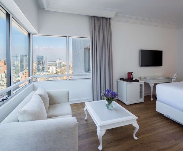 A modern hotel room with a large window offering a city view, a white sofa, a white coffee table with a vase, a small white table with a red object, a white bed, and a flat-screen TV on the wall. - NH Collection Plaza Santiago (Santiago)