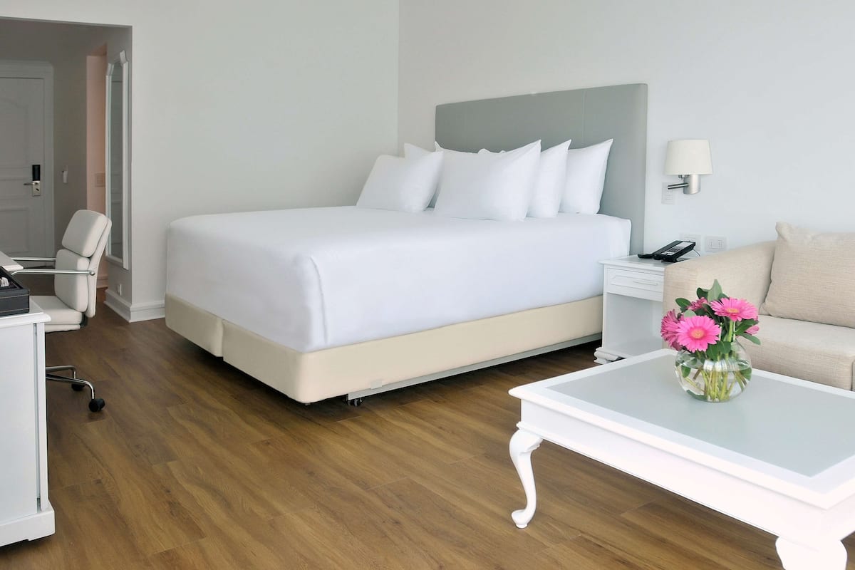 A hotel room with a large bed, a bedside table with a lamp, a small desk, a chair, and a coffee table with flowers.