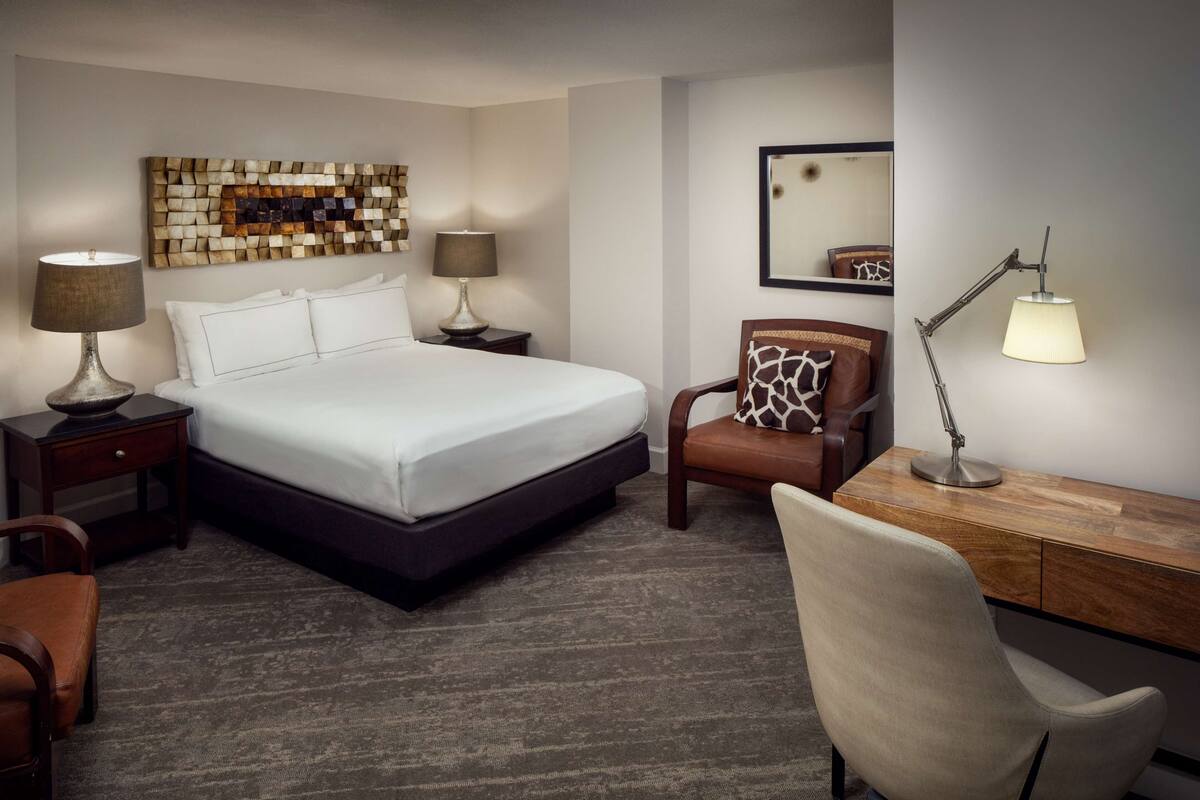A hotel room with a large bed, two bedside tables with lamps, a desk with a chair, and a wall art piece.