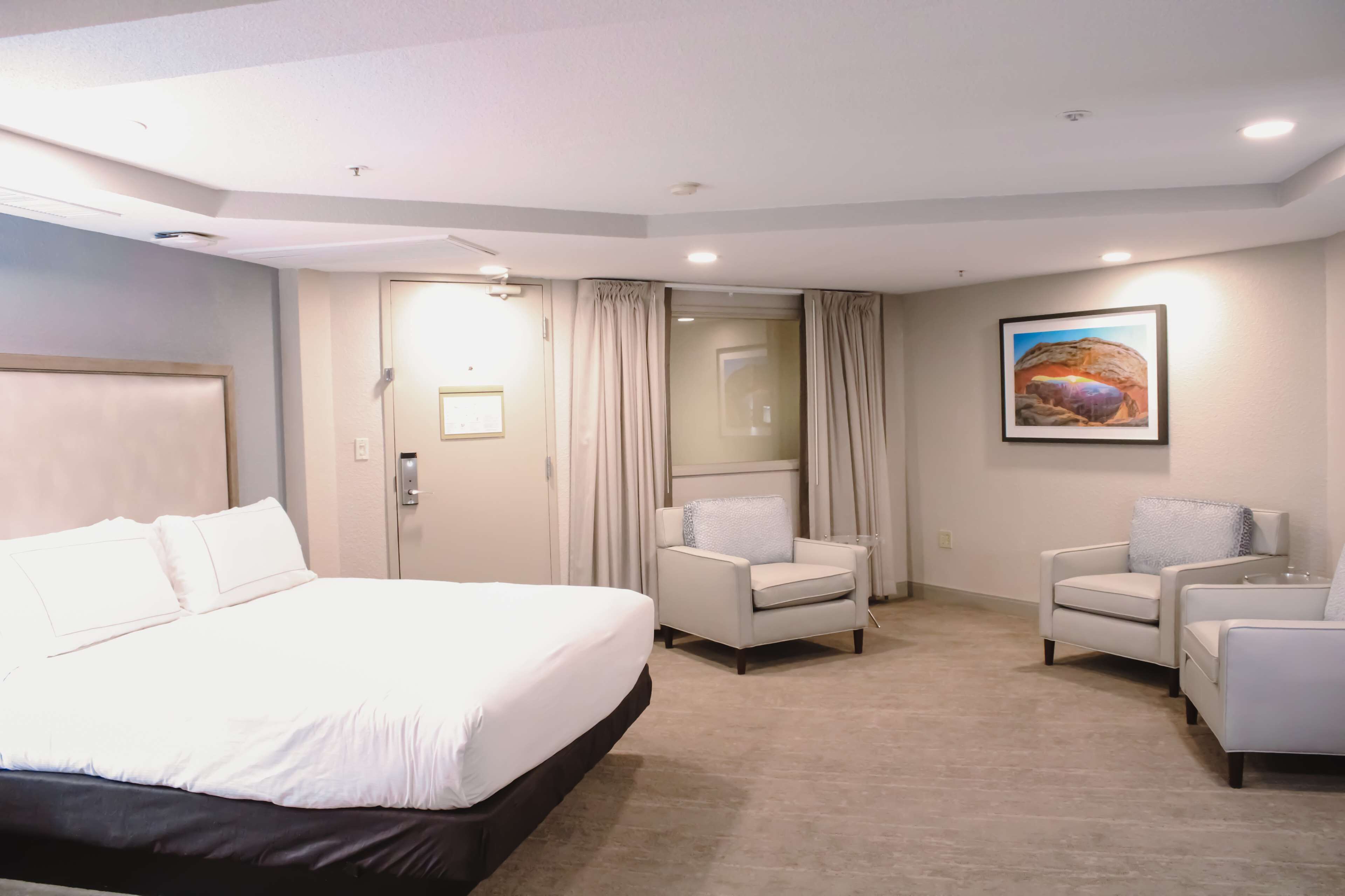 A hotel room with a large bed, two armchairs, a small table, a mirror, and a painting on the wall.