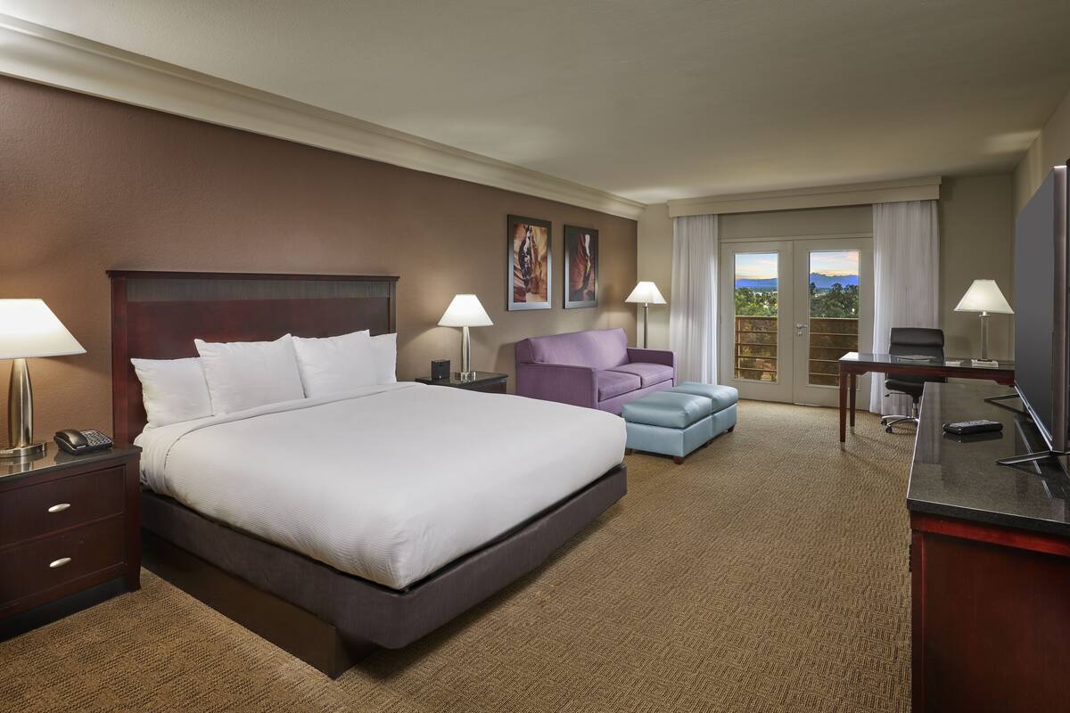 A hotel room with a large bed, two bedside tables, a desk, a chair, a purple armchair, and a view of the outdoors.
