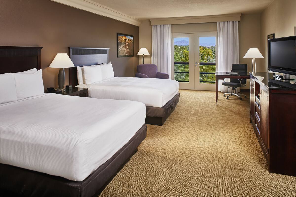 A hotel room with two beds, a desk with a TV, and a balcony with a view.