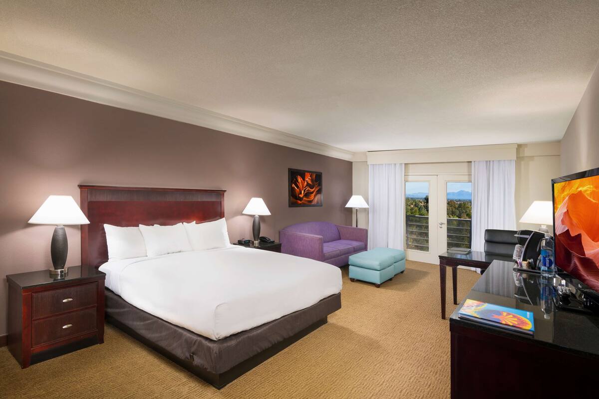 A hotel room with a large bed, two bedside tables with lamps, a purple armchair, a small blue ottoman, a desk with a TV, and a window with curtains.
