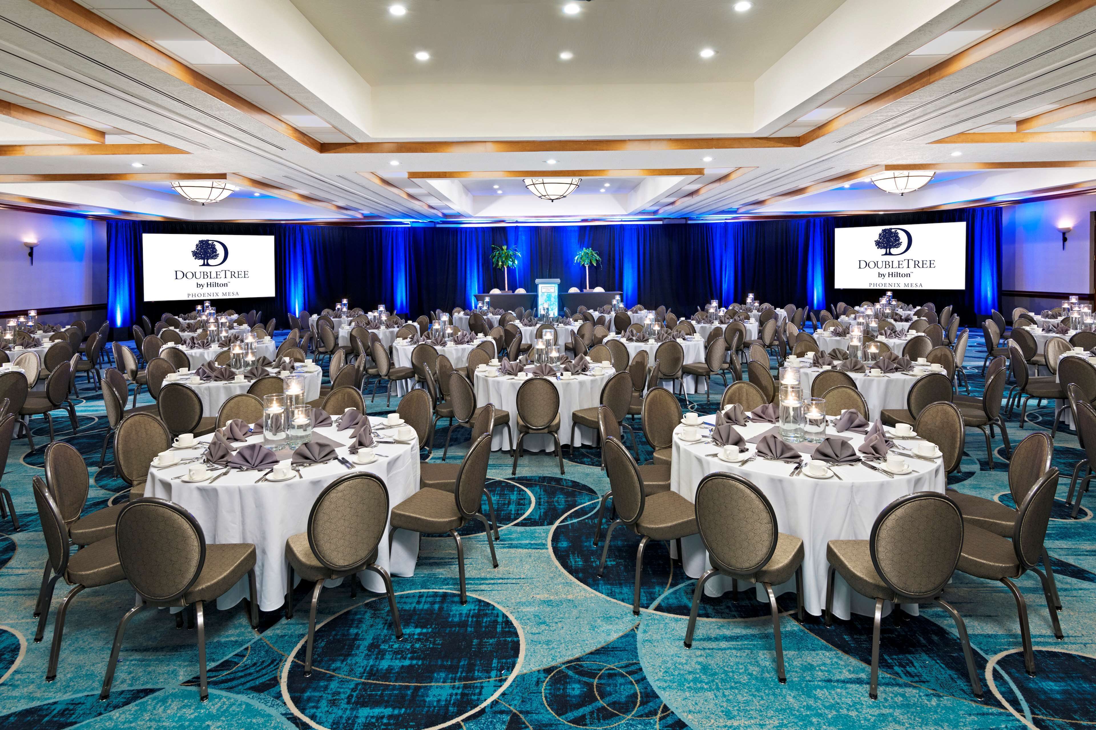 A large conference room with round tables and chairs, a stage with a podium, and a large screen displaying the DoubleTree by Hilton logo.