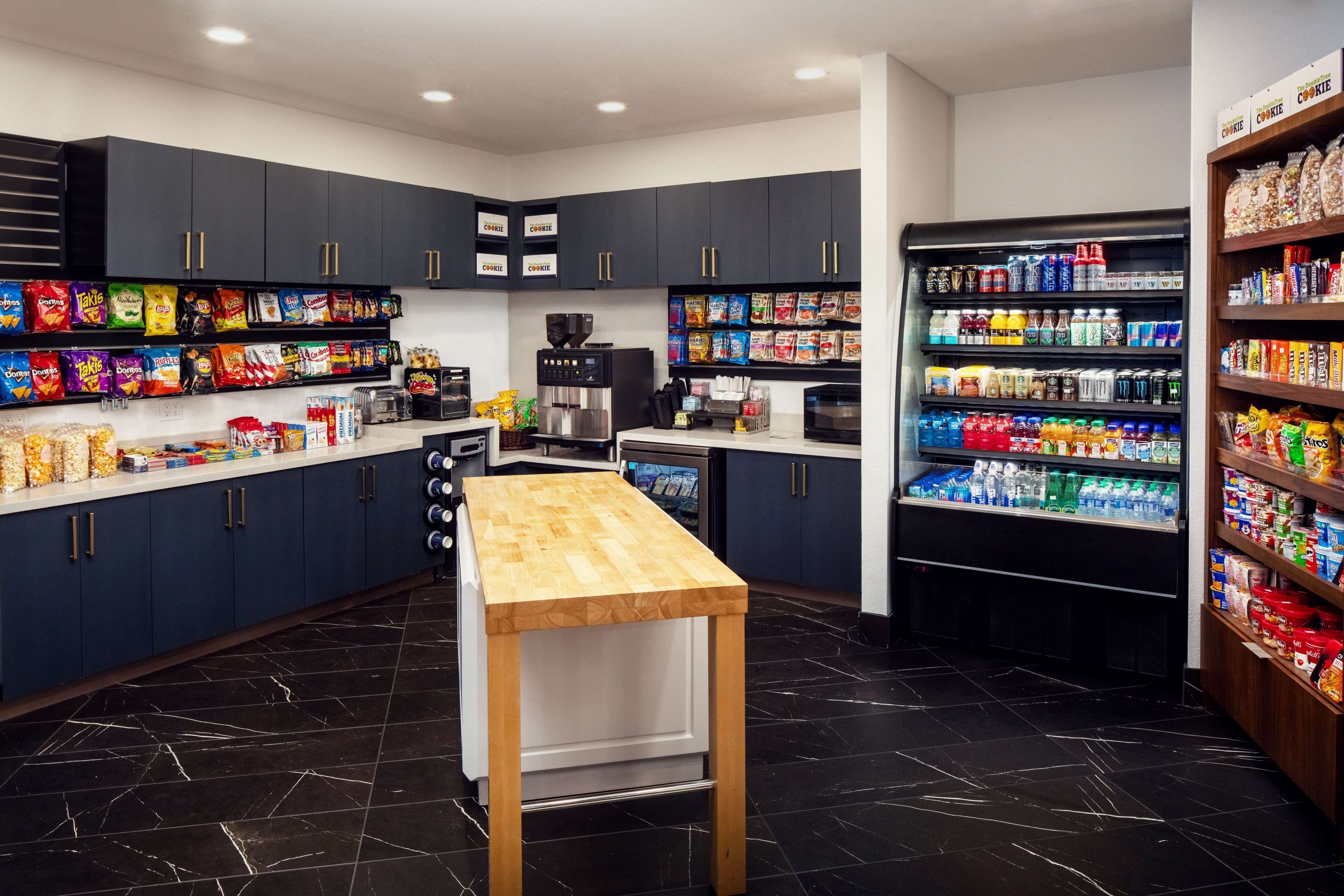 A modern kitchen with dark cabinets, a wooden table, and various snacks and beverages on shelves.