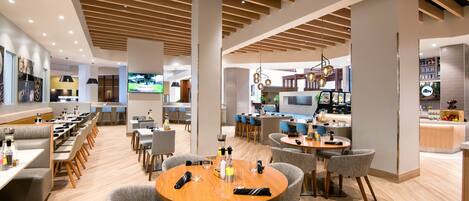 A modern hotel lobby with a central seating area, a bar, and a kitchen visible in the background.