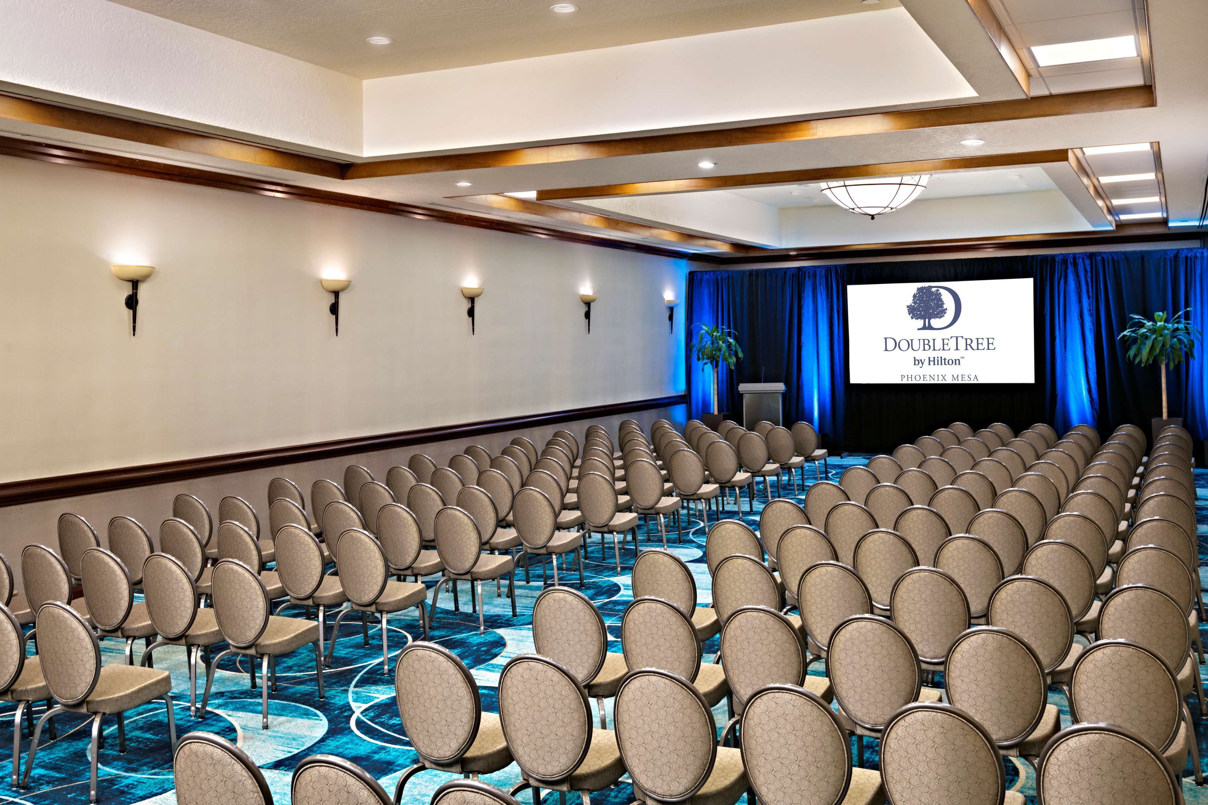 Une salle de conférence avec des rangées de chaises, une scène sur laquelle est affiché le logo « DOUBLETREE by Hilton » sur un écran, et un fond bleu en forme de rideau.