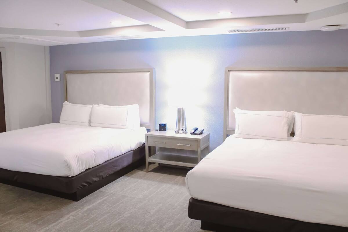 A hotel room with two beds, a nightstand with a lamp, and a window with blinds.